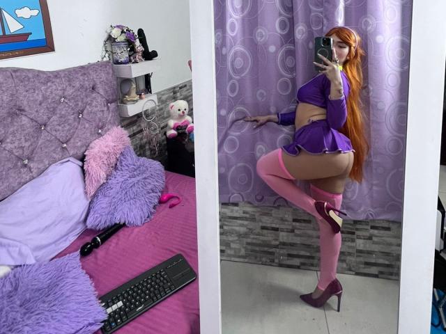 MelannyTaylorr with long orange hair in purple lingerie, showing her ass, in pink thigh-highs and heels, taking a mirror selfie, standing on one leg, in a bedroom.
