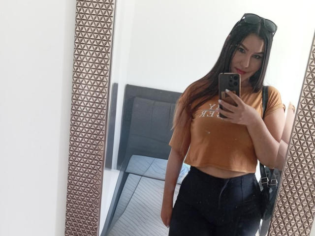 MariaRowe with long dark hair, orange crop top, black high-waisted pants, holding phone in mirror, sunglasses on head, small breasts, slightly visible ass, mirror with geometric frame.