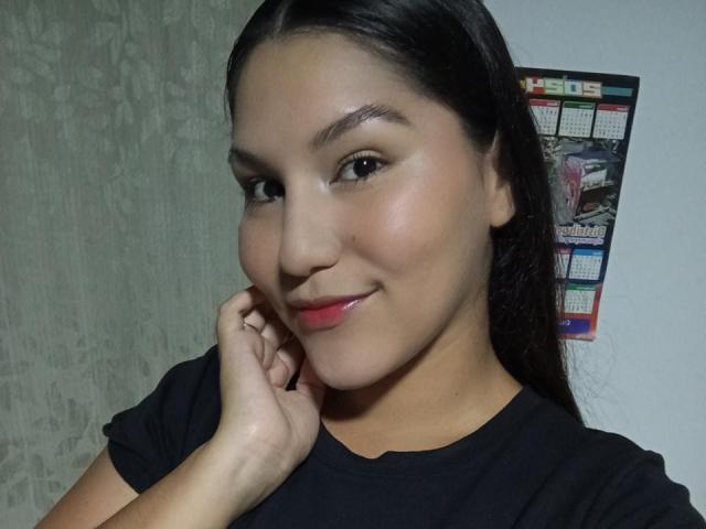 MariaRowe has dark hair, light brown skin, and red lipstick. She's wearing a black shirt, smiling slightly, and touching her cheek with her right hand.