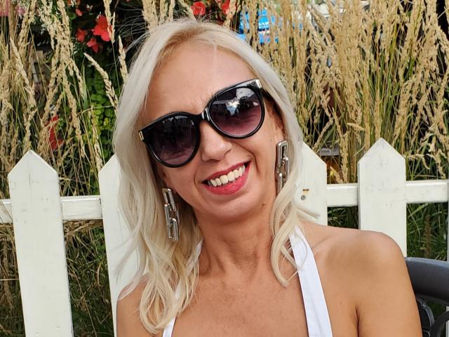 LunaBriella has blonde hair, wears black sunglasses, and a white halter top, smiling with red lipstick, showing her tits, and has large silver earrings, standing by a white fence.