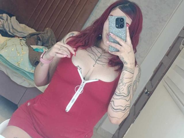 SaraJoyce with red hair, wearing a tight red dress with a white zipper, showing off her large tits and tattoos, taking a mirror selfie with a glittery phone.