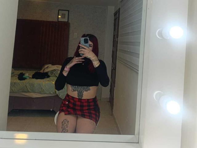 SaraJoyce with red hair, black long-sleeve top, red plaid mini-skirt, tattoos on stomach and thigh, mirror selfie, holding phone, bed and green blanket in background.