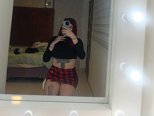 SaraJoyce with red hair, black crop top, red plaid skirt, tattoos on belly and thigh, taking mirror selfie, showing her ass and small tits, in dimly lit room.