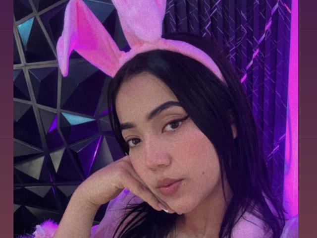 Ivone Sanz, con orecchie a coniglio rosa, capelli neri e lisci e eyeliner rosa, appoggia la guancia sulla mano, appare sensuale, con una luce al neon viola sullo sfondo..