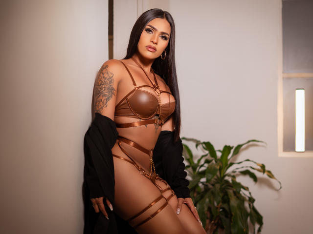 MadissonRush with long dark hair, brown leather strappy lingerie, black jacket off, tattoo on right arm, standing, looking at camera, potted plant in background, soft lighting.