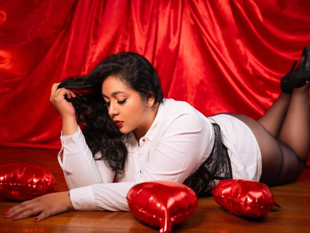ViickyLee, with curvy ass and dark skin, lies on wooden floor, wearing white shirt, black lace, and black heels, touching black hair, with red heart pillows around.