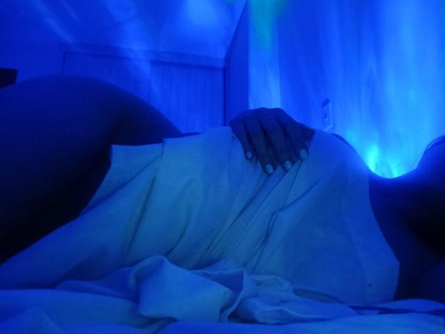 DanaLamiri lies in a blue-tinted bed, wrapped in a white towel, hand on her tits, legs slightly spread, visible ass on the left.