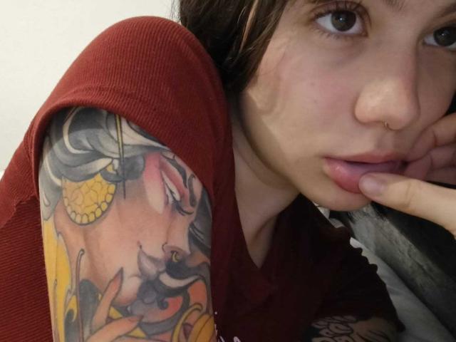 IsabellaArias with a nose ring, brown eyes, and slightly parted lips, wearing a red shirt, showing a colorful tattoo of a woman with a yellow flower on her shoulder.