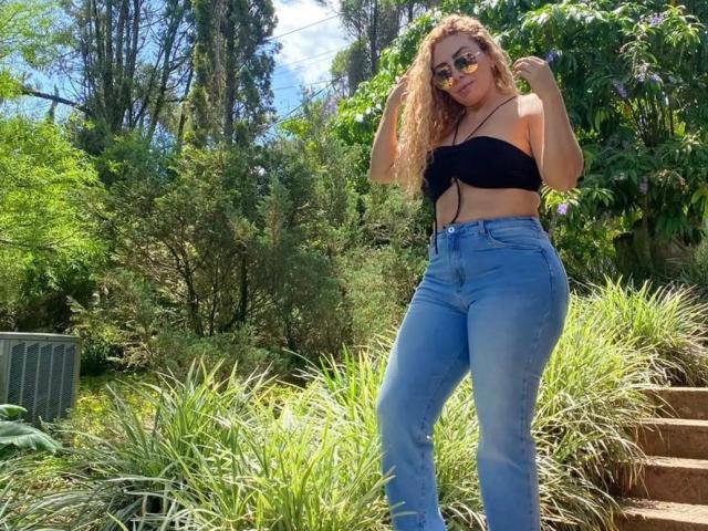 NaniDelaPavva, con cabello rubio rizado, vestida con un top negro sin tirantes, jeans azules de cintura alta y gafas de sol, está de pie en un jardín soleado con vegetación y escaleras. Sus pechos son pequeños..