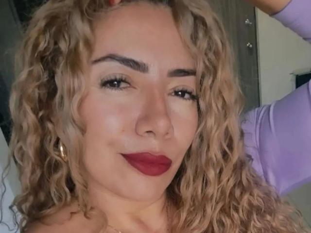 NaniDelaPavva, con i capelli biondi ricci, il rossetto rosso e un leggero sorriso, indossa una camicia viola, ha un piercing al naso e orecchini dorati, e appare sicura di sé..