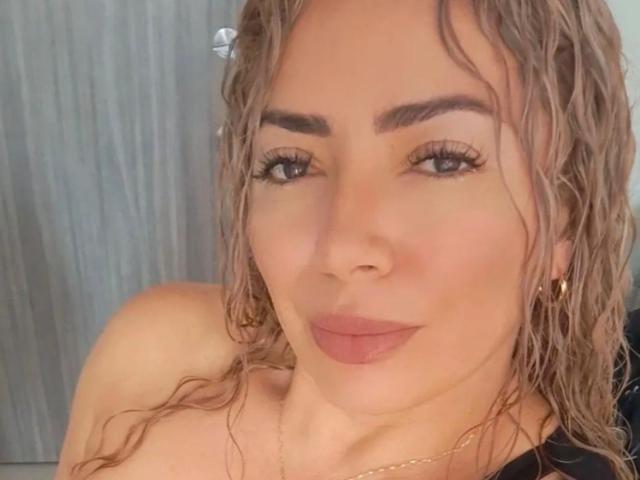 NaniDelaPavva tem cabelos loiros cacheados e úmidos, sobrancelhas escuras, cílios longos e está fazendo uma expressão de lábios emburrados com batom rosa. Ela está usando brincos de argola dourados e uma blusa preta..