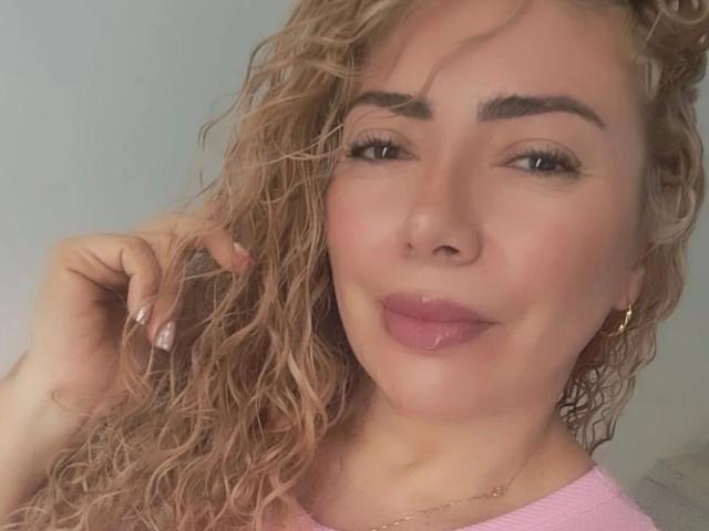 NaniDelaPavva, con i capelli biondi ricci, le sopracciglia folte, il rossetto rosa, gli orecchini dorati e una maglietta rosa, sorride dolcemente, toccandosi i capelli con una mano con le unghie dipinte di rosa..
