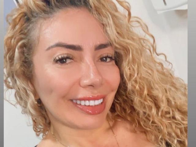 NaniDelaPavva hat lockiges, blondes Haar, braune Augen, dicke Augenbrauen und einen glänzenden, roten Lippenstift. Sie lächelt und zeigt ihre weißen Zähne. Ihre Nase ist klein und sie trägt eine Halskette..