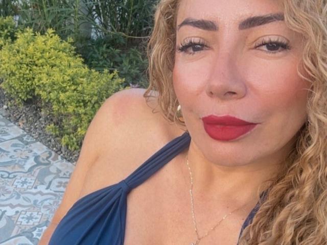 NaniDelaPavva mit lockigem, blondem Haar, einem dunkelblauen Top mit Halter, rotem Lippenstift und einer goldenen Halskette, zeigt ein leichtes Lächeln, wobei ihre Brüste teilweise sichtbar sind, in einer sonnigen, offenen Umgebung..