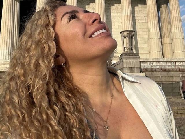 NaniDelaPavva, con i capelli biondi ricci, sorride guardando verso l'alto, indossa una camicia bianca, una collana con una croce, ha la pelle olivastra, con una scollatura visibile, ed è in piedi di fronte a colonne alte. La luce del sole illumina il suo viso..