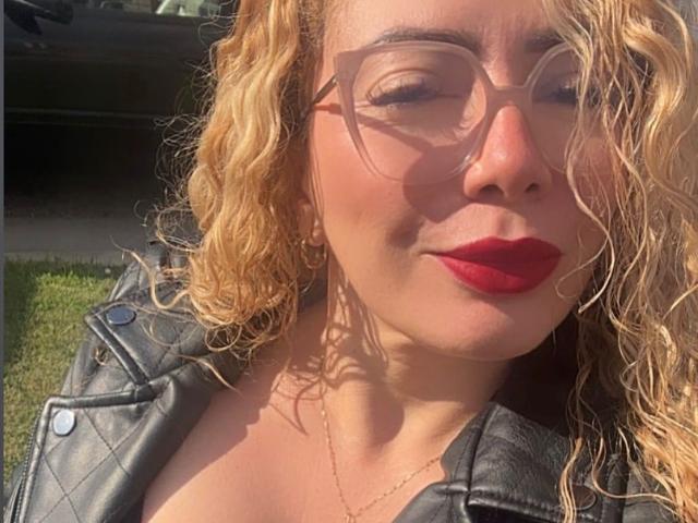 NaniDelaPavva mit lockigem, blondem Haar, rotem Lippenstift, Brille und einer schwarzen Lederjacke, lächelnd im Freien. Brüste sind unter der Jacke sichtbar, Sonnenlicht auf dem Gesicht..