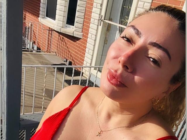 NaniDelaPavva has puffy lips, red tank top, and a cross necklace, with light brown hair in a ponytail, on a sunlit balcony. Her eyes are half-closed, looking cute.