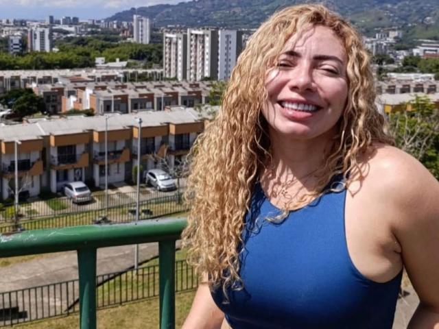 NaniDelaPavva, avec ses cheveux blonds bouclés, souriante, portant un haut de sport bleu, montrant ses seins, debout près d'une rambarde verte, avec une ville et des montagnes en arrière-plan, une journée ensoleillée..
