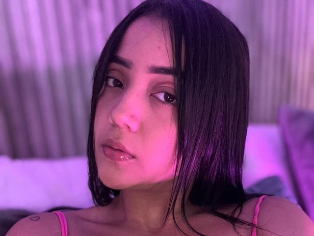 Ivone Sanz, com o cabelo preto molhado, iluminação rosa no rosto e uma pequena tatuagem no ombro esquerdo, com uma expressão ligeiramente entediada e os lábios rosados entreabertos. Seios não visíveis..