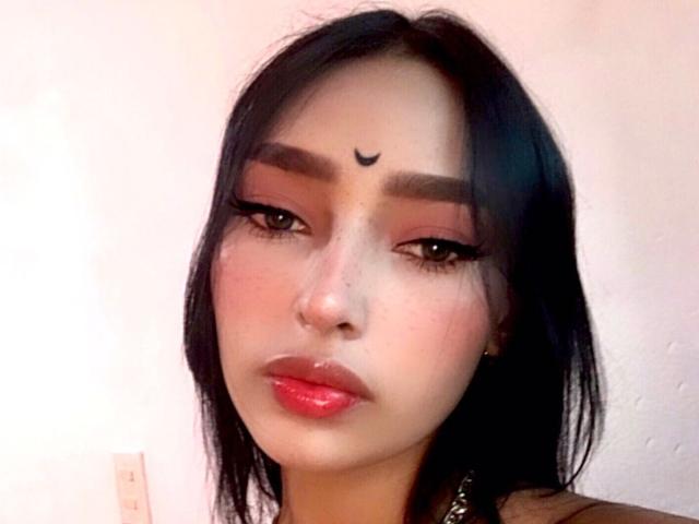 AdelaBanks has straight black hair, parted slightly to the side, with a small crescent-shaped tattoo above her forehead. She has dark, thick eyebrows, long eyelashes, and red lipstick. Her eyes are slightly downcast, giving a pensive look. She wears small hoop earrings and a black strap is visible on her right shoulder. The skin tone is light with a subtle pinkish hue. The image has a soft, almost pastel-like quality. The focus is on AdelaBanks's face, which is centered and takes up most of the frame.