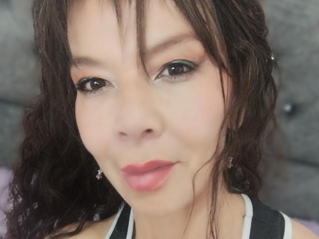 SamaraBellet has wavy brown hair, green eyeshadow, pink lipstick, and small earrings. Her face is slightly blurred with a soft smile, showing light skin and subtle makeup.
