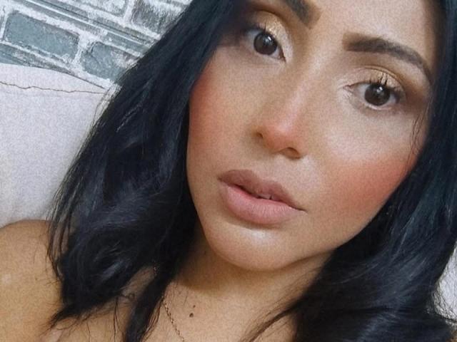 Het gezicht van Taliana Prada, met donker golvend haar, bruine ogen en lichtroze lippen, toont een subtiele, aantrekkelijke uitdrukking. Haar lichtbruine huid is glad en ze kijkt naar de camera..