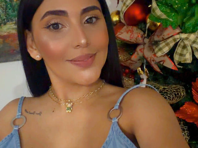 Taliana Prada, avec ses cheveux foncés, ses yeux marron et son sourire discret, porte un haut bleu avec des détails en forme d'anneaux, un collier en or et des boucles d'oreilles, et on peut apercevoir un petit tatouage sur son épaule gauche..