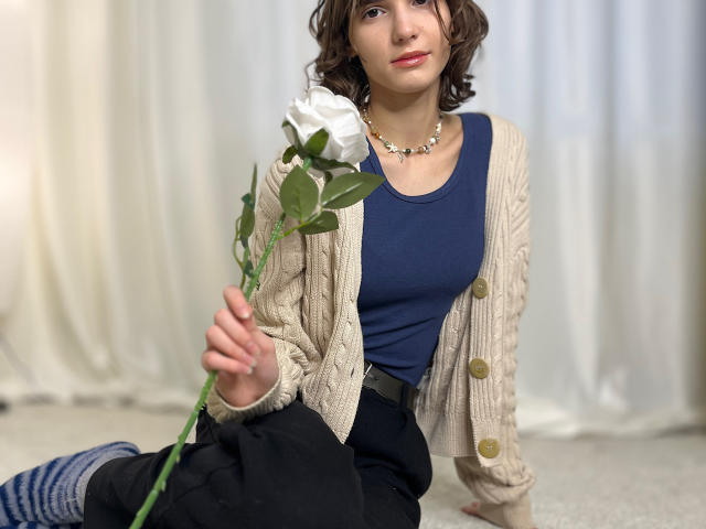 ErynAttridge sits on a carpeted floor, holding a white rose. She has short, curly brown hair and wears a navy blue shirt, black pants, and a beige knitted cardigan with large buttons. She also has a necklace with small, clear stones. Her right hand holds the rose's green stem, and she sits with her left arm slightly behind her. Her blue and white striped socks are visible. The background is a plain white curtain. The image is a photograph.