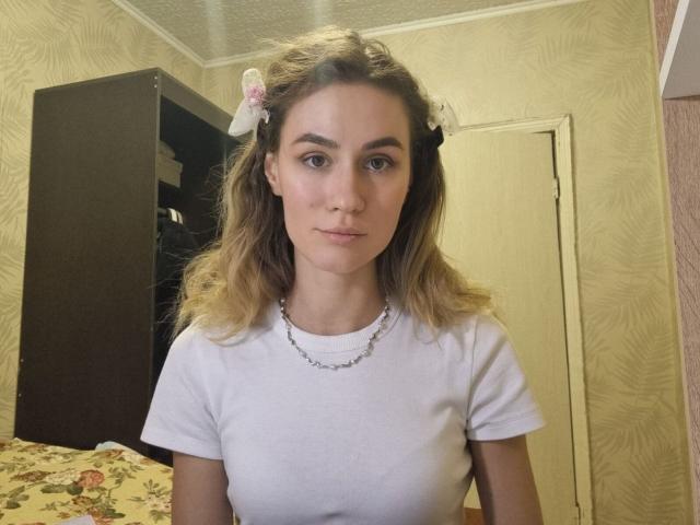 LorenaPenix has blonde hair, white shirt, small tits, and a neutral expression. She's wearing a floral hair clip and a chain necklace in a modestly decorated room.