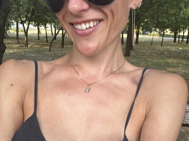 BellaJulls with a smile, wearing black straps, sunglasses, and a necklace with a small pendant, showing her tits and a cute nose. She's outside with trees.