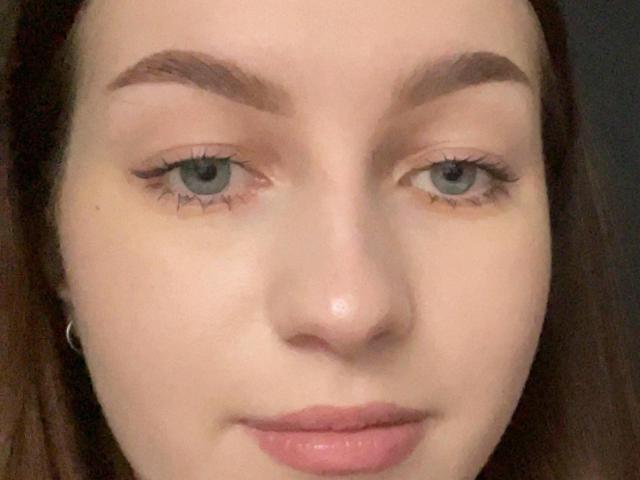 Le visage de Sara Rosenbarger, montrant sa peau pâle, ses yeux bleus, ses sourcils épais et bruns, ses longs cils, ses lèvres roses et ses cheveux bruns, avec un léger sourire. Ses seins ne sont pas visibles..