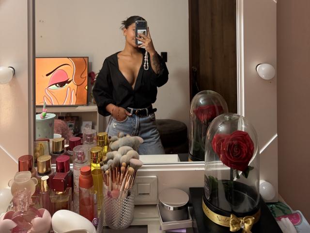 ThaliaAres, avec sa peau brune et ses cheveux foncés, portant un haut noir à décolleté plongeant qui révèle ses seins, un jean bleu taille haute, tenant un téléphone, debout devant un miroir rempli de produits de maquillage..