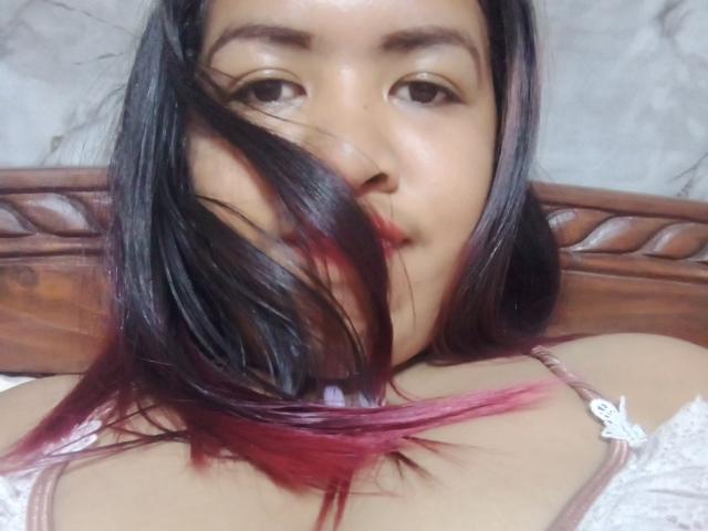 MariahSquirting69 with dark black and pink-tipped hair, slightly parted lips, and brown eyes, lying on a wooden headboard, wearing a white lace top, showing her bare shoulders.