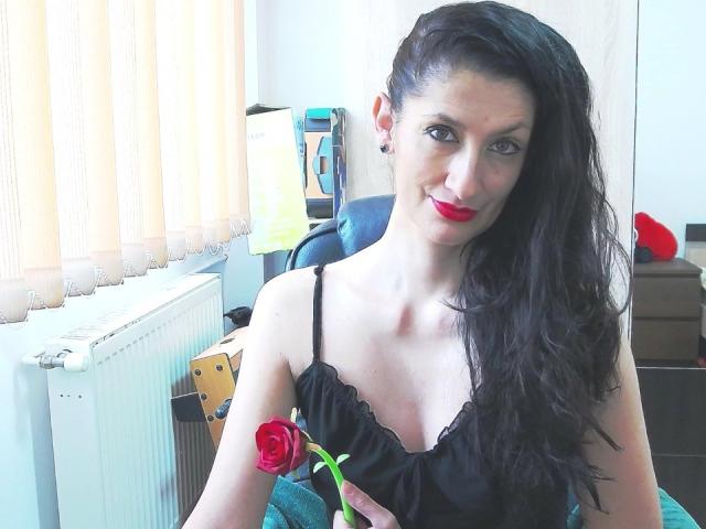 BeatrixCharmm with long black hair, red lipstick, black spaghetti top, holding a red rose, sitting in a room, white radiator, and yellow light on a desk behind.