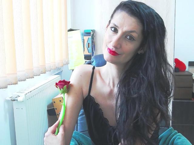 BeatrixCharmm with long black hair, red lipstick, and black lace lingerie, holding a red rose, sitting near a window, and smiling at the camera.