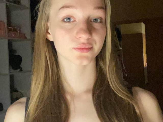 ArielleFiorello has pale skin, blue eyes, and long blonde hair. She's wearing a black tank top, showing her small, bare tits. She's looking at the camera with a slight smile.