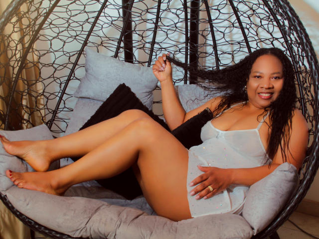 SharonNoa with curly black hair, wearing a sheer white dress, showing her ass and tits, lying in a black hanging chair, smiling, holding her hair.