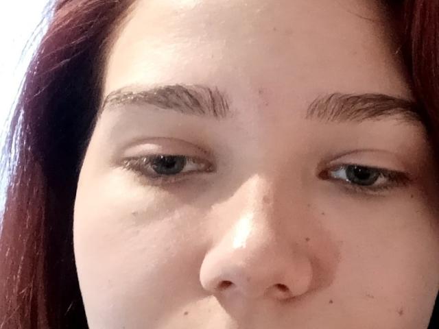 GerdaCastino's face, showing pale skin, blue eyes, and dark red hair. Her eyebrows are thick and she has small freckles and moles. Her nose is centered in the frame.