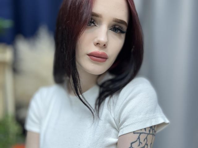 AleneNghiem a les cheveux roux, porte une chemise blanche et a un tatouage sur le bras droit. Elle regarde vers le bas, avec des lèvres roses pincées, un trait d'eye-liner noir et un anneau au nez. Ses seins sont petits et plats..