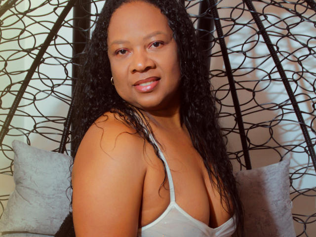 SharonNoa with wet black curly hair, medium brown skin, small gold hoop earring, and a white tank top showing her tits, smiling slightly with pink glossy lips.