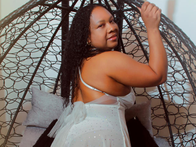 SharonNoa with curly black hair, wearing a white sparkly dress, sitting in a black wicker egg chair, looking back with a smile, showing her back and small round butt.