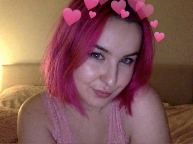 Lionelly with pink hair, cute smile, and pink lace top, has hearts over her head, sitting on a bed, looking at the camera, showing her bare shoulders.