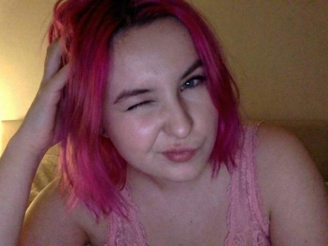 Lionelly with pink hair, winking one eye, hand in hair, wearing a pink lace top, has a slight smirk and pale skin, looking cute and slightly mischievous.