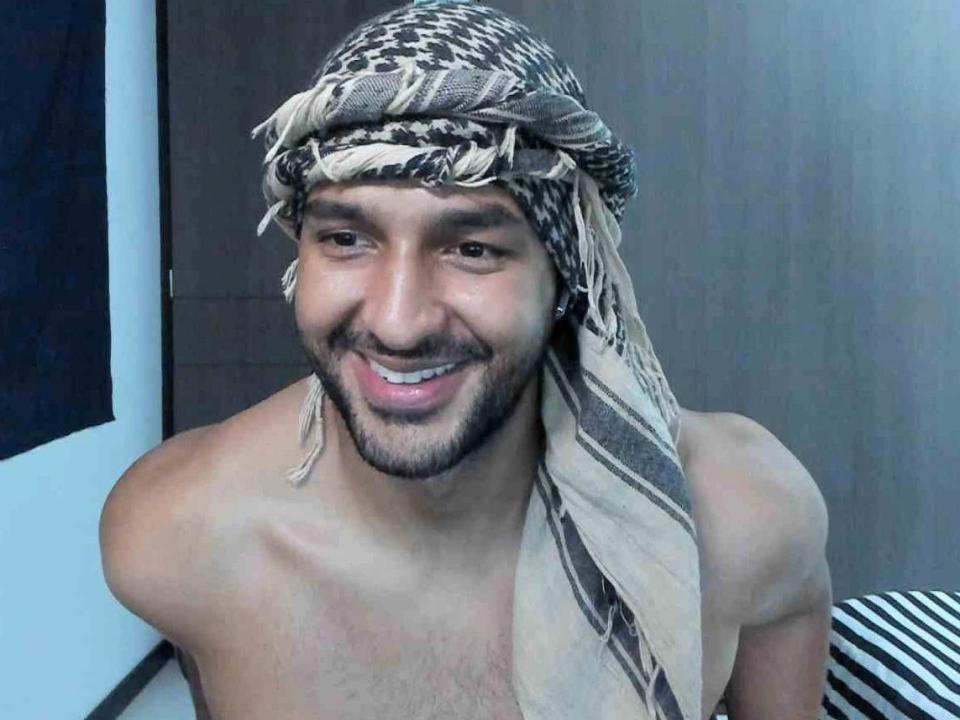 RalfVoss is een man zonder shirt, met een baard, die lacht en een gekleurde hoofddoek draagt. Hij heeft een gemiddeld postuur en een bruine huid. Zijn achterkant is niet zichtbaar..