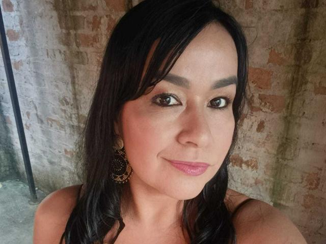 TamaraFoxy with dark hair, brown eyes, pink lipstick, and black earrings, smiling slightly, showing her face and upper shoulders. She has medium-sized tits and a cute look.