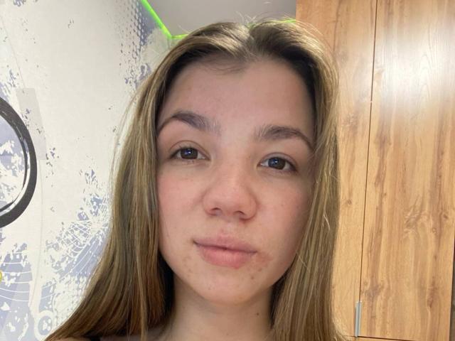 Marilynn Mcmasters hat hellbraunes Haar, blaue Augen und Sommersprossen im Gesicht. Sie schaut direkt in die Kamera und lächelt leicht. Ihre Nase ist klein und niedlich..
