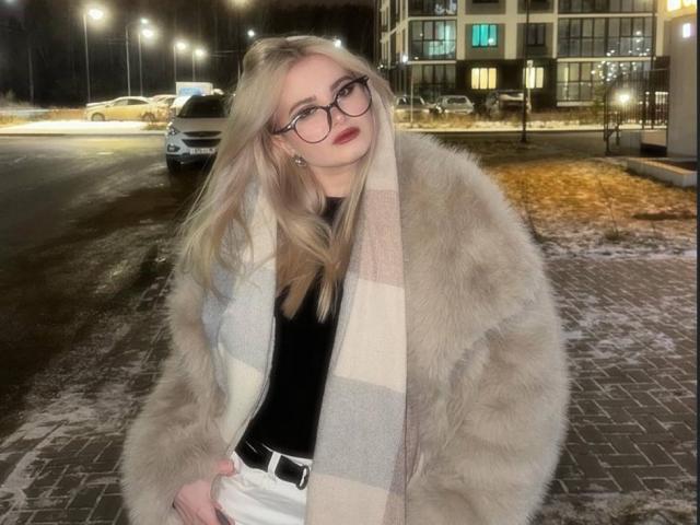 Nichelle Tetro, com cabelo loiro, óculos e batom vermelho, vestindo um casaco de pele cinza fofo e calças brancas, em um cenário noturno urbano com neve..