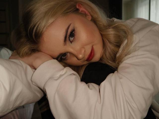 Nichelle Tetro met blond haar, rode lippenstift en een wit shirt met lange mouwen, leunt haar hoofd op haar hand en kijkt omhoog met een lichte glimlach. Haar achterkant is gedeeltelijk zichtbaar..