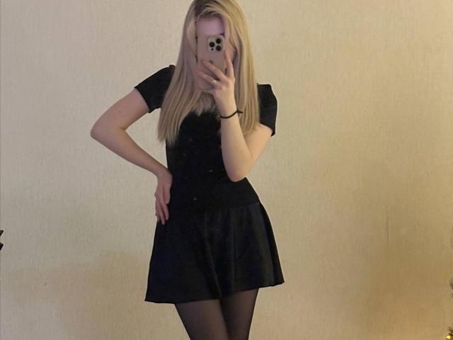 NichelleTetro con i capelli biondi, una maglietta nera a maniche corte, una gonna nera e collant neri, che tiene un telefono, coprendosi il viso, in piedi con una mano appoggiata al fianco..