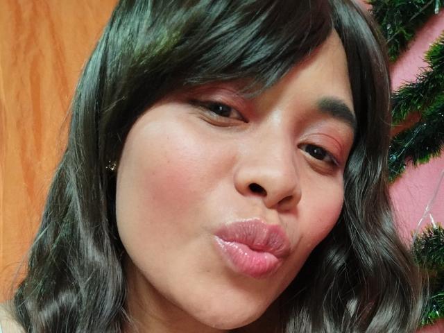 MithyMiss pouts her glossy pink lips, with dark wavy hair, and slightly flushed cheeks, looking into the camera with a subtle, confident expression. Her nose is centered.